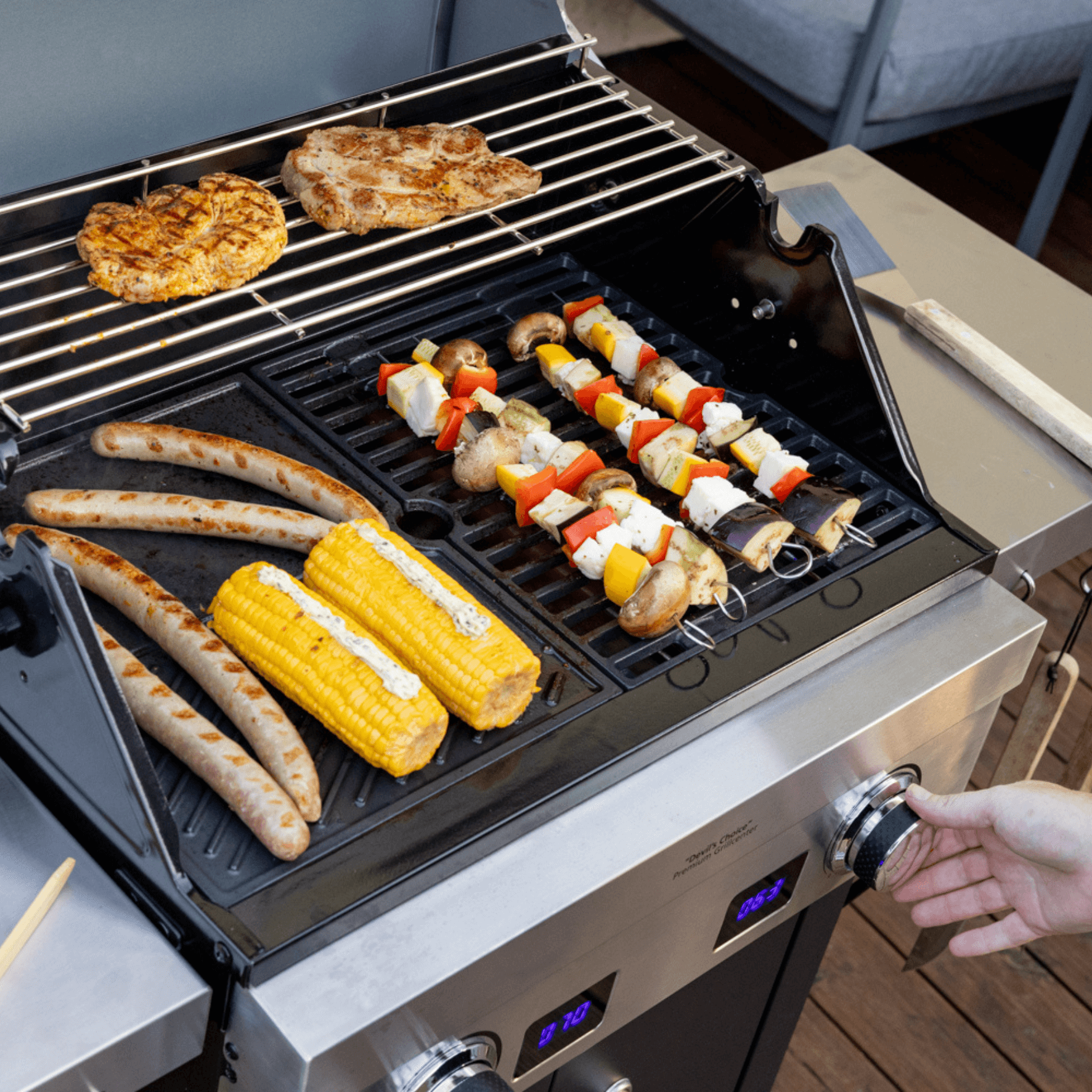 Steba VG7000 BBQ-Grill mit Gemüse- und Fleischspießen, Würstchen und Maiskolben auf dem Grillrost. Temperaturregler sichtbar.