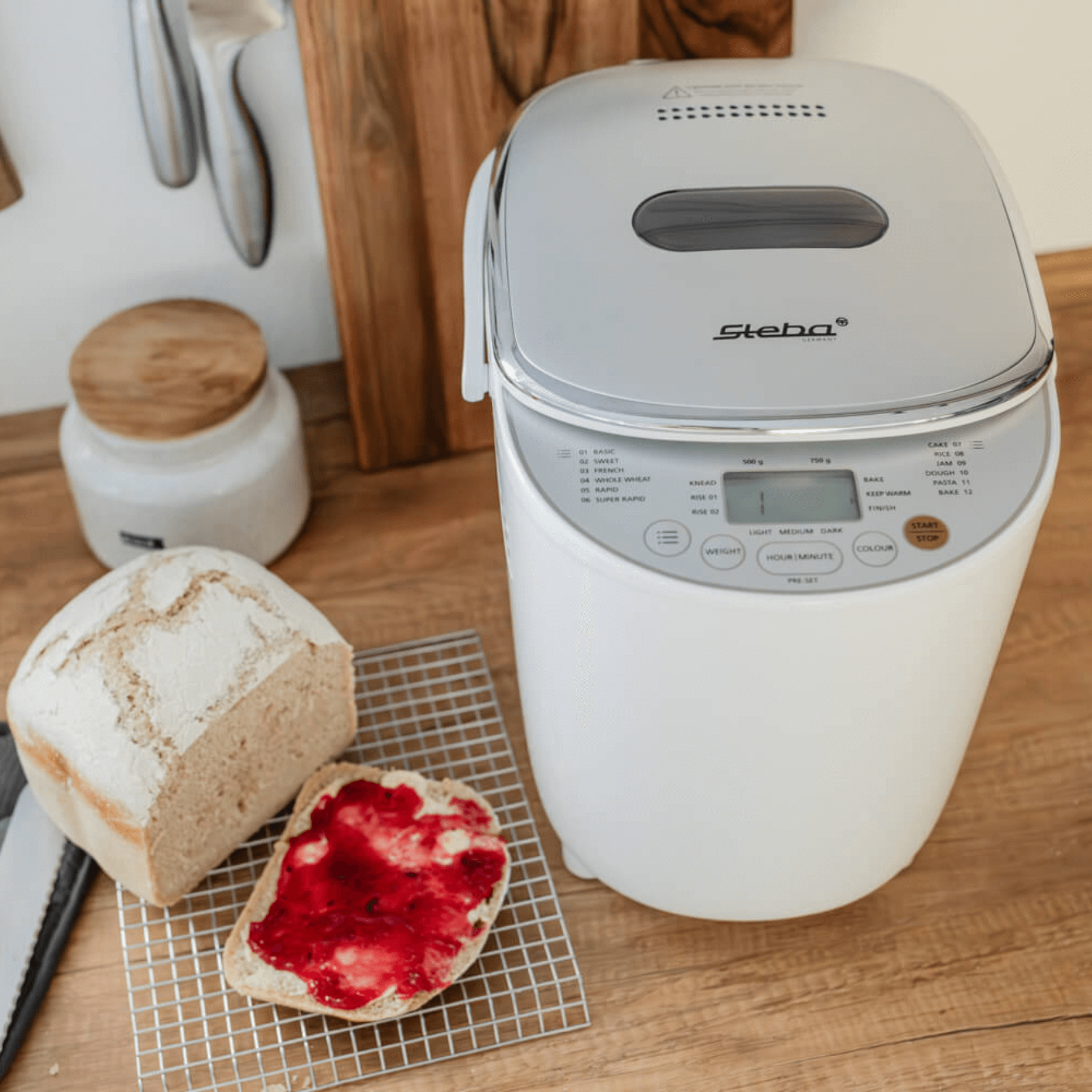 Steba BM3 Brotbackautomat in Weiß mit Bedienfeld. Neben dem Gerät liegt ein frisch gebackenes Brot und ein Toast mit Marmelade.