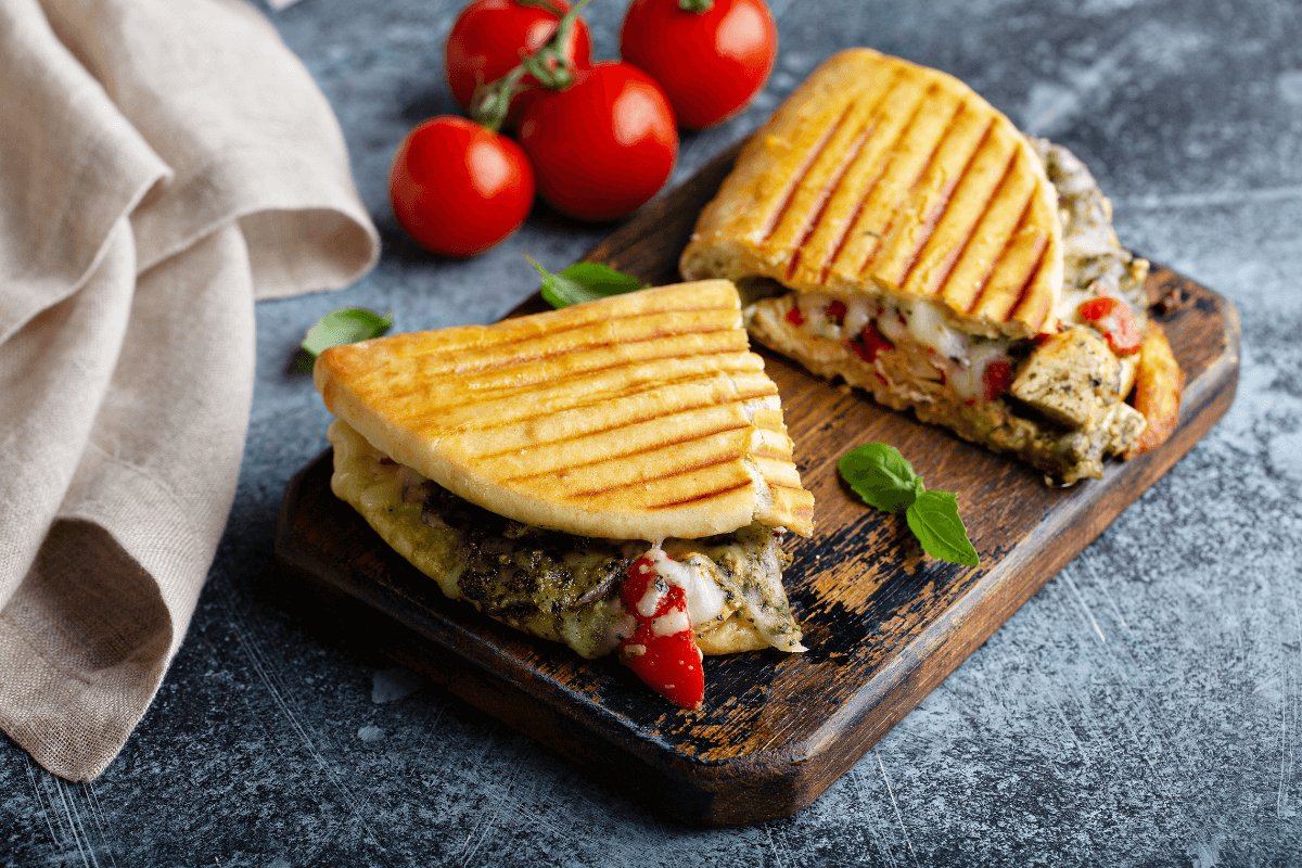Zwei gegrillte Panini mit Gemüsefüllung, serviert auf einem Holzbrett, daneben frische Tomaten und Basilikumblätter.