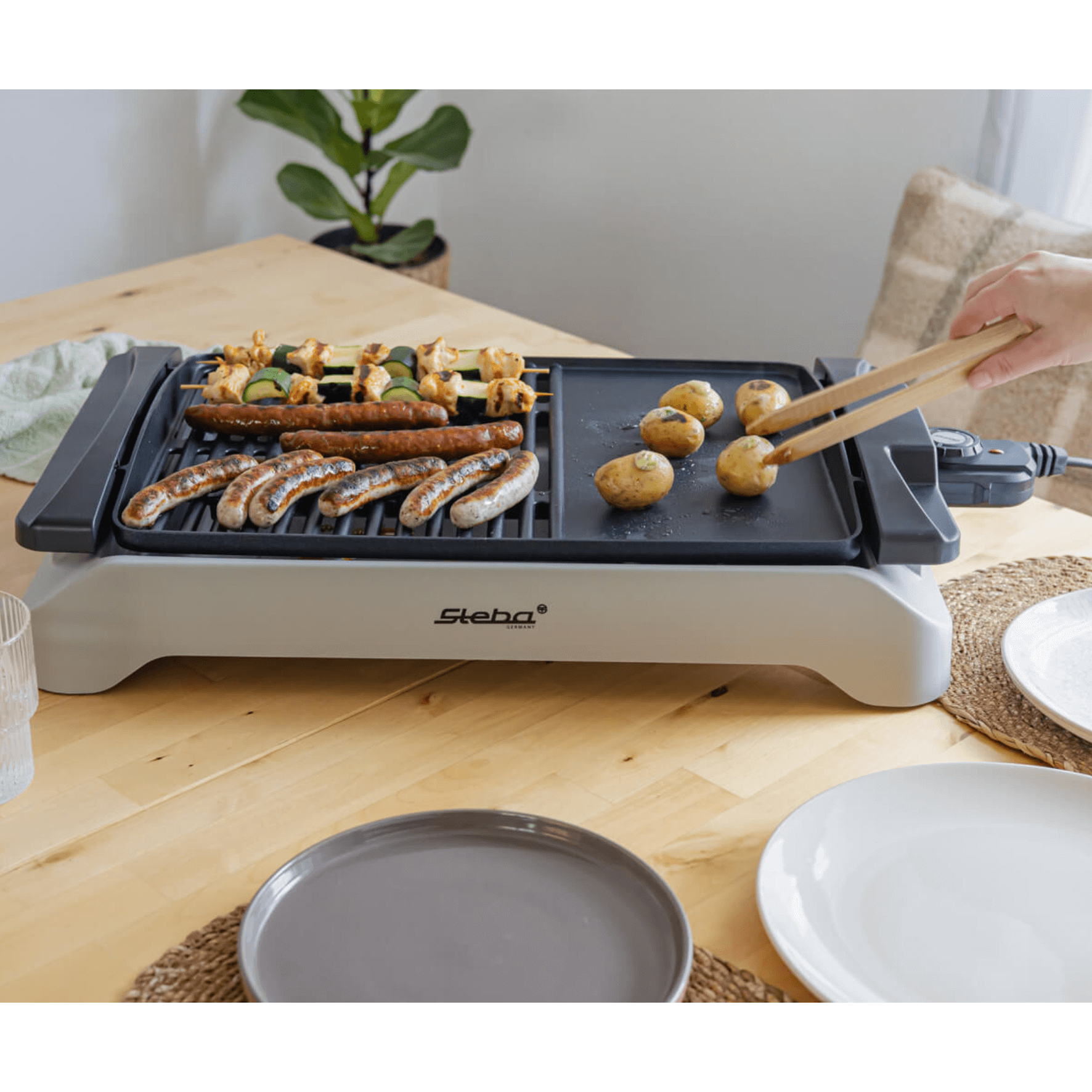 Steba VG101 BBQ-Grill mit Grillfläche und Pfanne, Fleisch und Gemüse in Zubereitung, auf einem Tisch mit Geschirr.