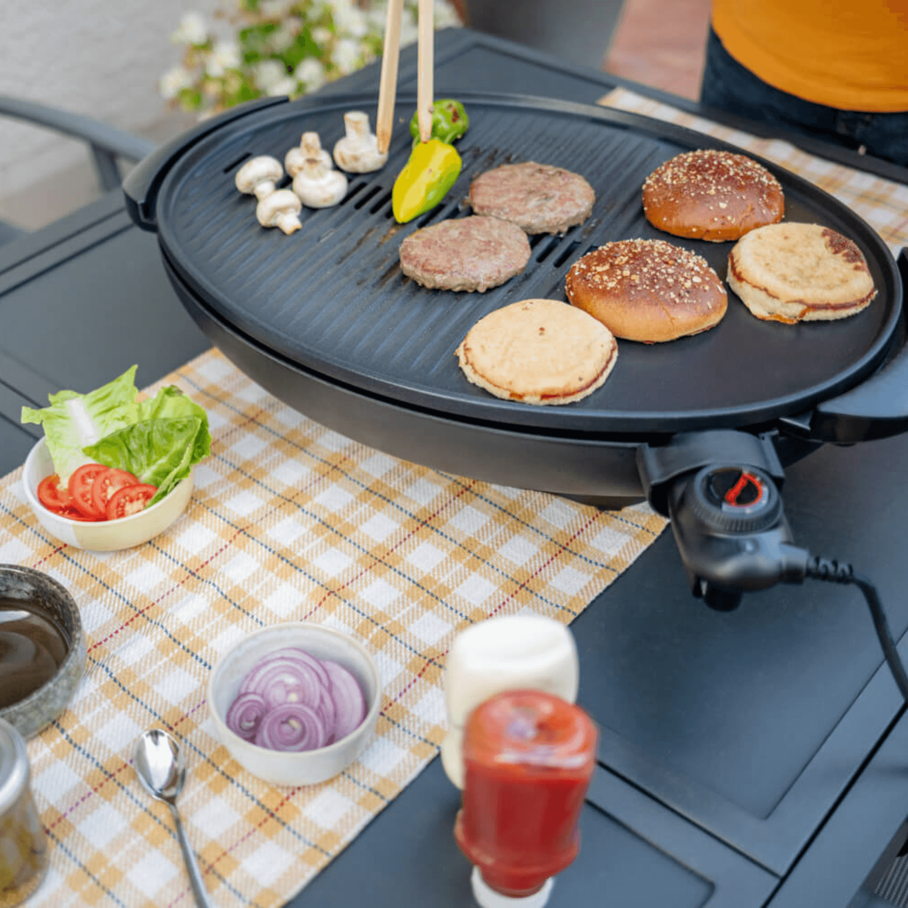 Elektrogrill Steba VG350 mit Grillfläche, auf der Burger, Brötchen und Gemüse zubereitet werden. Zubehör wie Soßen und Gemüse sind sichtbar.