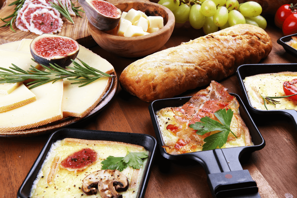 Raclette-Gerichte in Pfännchen mit Käse, Schinken, Pilzen und Tomaten, umgeben von Brot, Käse, Feigen und Trauben.