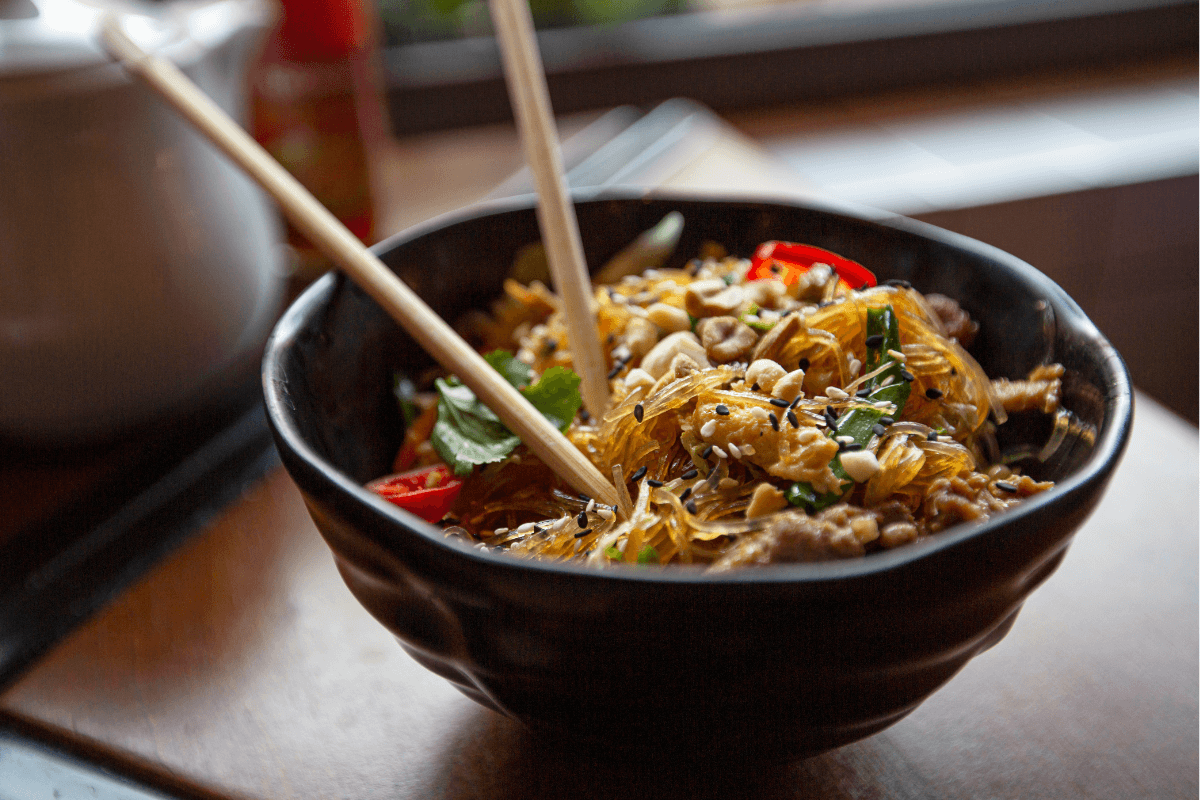 Schwarze Schüssel mit asiatischem Glasnudelsalat, garniert mit Kräutern, Erdnüssen und roten Chilis, serviert mit Essstäbchen.
