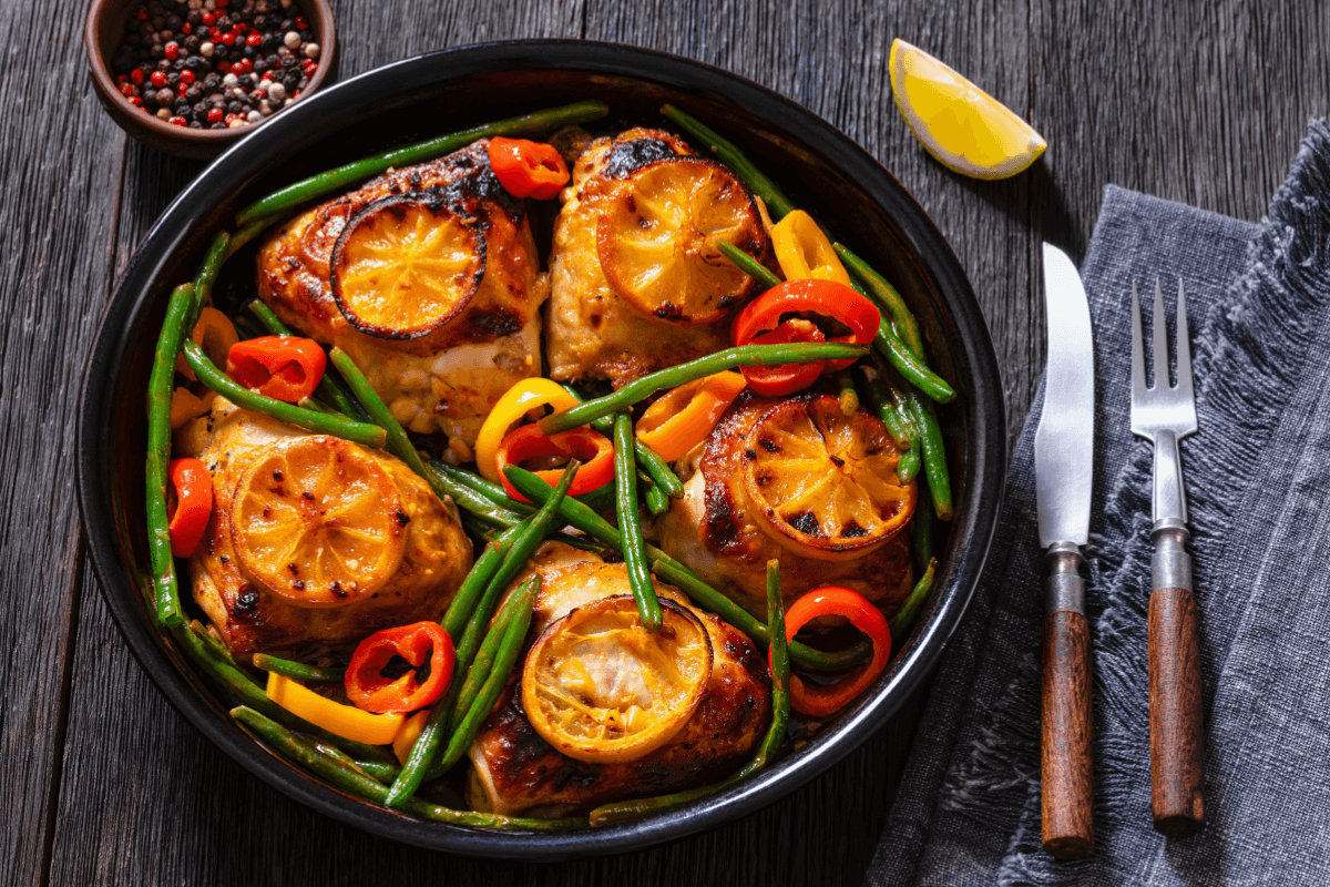 Zitronenhähnchen mit grünen Bohnen, Paprika und Zitronenscheiben in einer schwarzen Servierschüssel.
