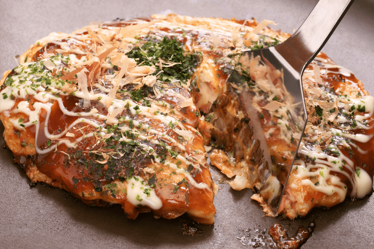 Okonomiyaki auf einer Grillplatte, garniert mit Mayonnaise, Sauce, Algen und Bonitoflocken. Ein Stück wird herausgehoben.