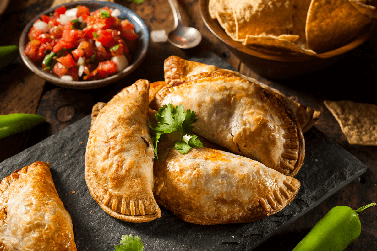 Krustige Empanadas auf einem schwarzen Steinbrett, garniert mit frischem Koriander, daneben Salsa und Tortilla-Chips.