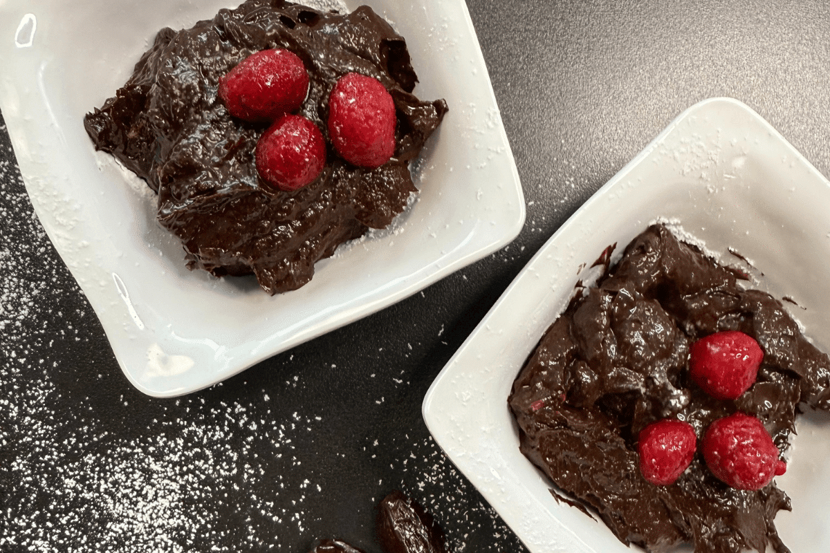 Vegane Mousse au Chocolat in weißen Schalen, garniert mit frischen Himbeeren und Puderzucker, auf schwarzem Untergrund.